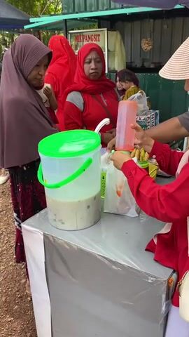 The video shows an outdoor scene with several people, likely at a market or food stall. In the foreground, a person wearing a red shirt and a white hat is handing a red plastic bottle to another person also in a red shirt. In front of them is a large plastic container with a green lid, filled with a light-colored liquid containing various colorful ingredients. To the left, two women wearing headscarves are standing. One woman, wearing a maroon headscarf and a patterned dress, is holding a child. Another woman in a bright red headscarf is looking towards the stall. In the background, there are tents, tables with food items, and more people, including children. The environment appears to be outdoors with dirt ground and trees visible. A sign with text is partially visible above the stall.