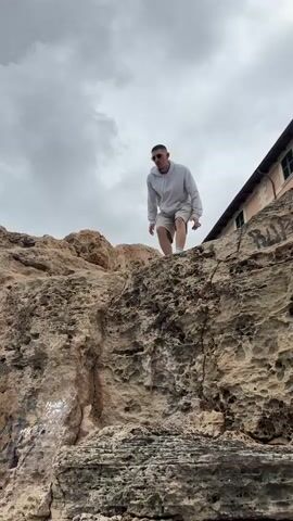 A young man in a white hoodie, light-colored shorts, and white shoes stands on a rocky outcropping overlooking the ocean. He has short hair and is wearing sunglasses. The rocks are a mix of browns and grays, and have some graffiti on them. The ocean is visible behind the rocks, and the sky is overcast with grey clouds. He sits down on the edge and another person appears in the foreground, taking a photo. In the background, there is a building with a reddish-brown roof and a port with several ships. The overall tone of the scene is casual and relaxed, despite the potentially dangerous location of the man.