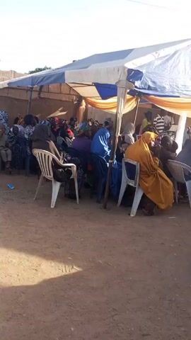 The video depicts an outdoor gathering under a tent. People are seated on chairs, listening to a speaker. A man wearing traditional white attire, a dark brown hat, and a beard looks directly at the camera. The tent is decorated with blue and gold fabric. The setting appears to be a yard with a dirt surface.
