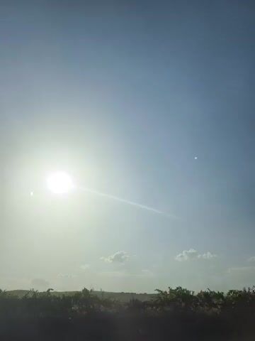 The video shows a daytime scene with the sun shining brightly in a clear blue sky. Power lines are visible in the upper portion of the frame, stretching across the sky. Below the power lines, a landscape with trees and vegetation can be seen. The sun is positioned in the left part of the frame, casting a strong glare. In the lower right corner, there's a glimpse of what appears to be a road or path, with some dust or debris being kicked up, suggesting movement. The overall impression is of an outdoor environment, possibly a rural or semi-rural setting, on a sunny day.