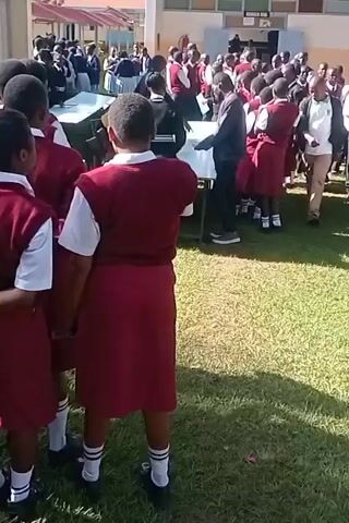 The video shows a group of students in maroon and white school uniforms gathered outdoors on a grassy area. They appear to be waiting or observing something happening in front of a building with a sign that is not clearly legible. Some students are standing by tables. A woman in a white shirt, black pants, glasses and a maroon scarf walks past the camera, carrying a brown envelope or folder. She is also wearing a badge. The environment suggests a school setting.