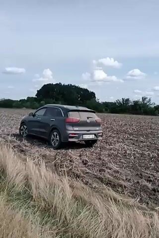 A dark gray Kia Niro is driving through a plowed field. The car is mostly seen from the rear passenger side. The field appears to be dry and dusty, with some remaining stubble from a previous crop. Trees are visible in the background. The sky is partly cloudy. The video is shaky, suggesting it was recorded by someone holding a mobile device. The car's license plate is visible and appears to be Ukrainian.