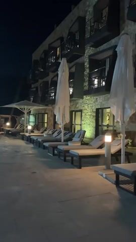 A dimly lit outdoor scene shows a row of empty lounge chairs lined up next to a stone-clad building at night. The chairs are made of wood and have gray cushions. Several closed white umbrellas are positioned among the chairs. The building has multiple floors with balconies and windows that are illuminated from within. The ground is paved with stone tiles. Small, modern outdoor lamps are placed along the walkway.