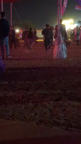 The video shows a group of people at night, standing on a sandy or muddy surface. There are several individuals walking around. Some are in short skirts or shorts, while others are in jeans or long pants. A man in a dark jacket is standing to the left of the frame, facing away from the camera. In the middle of the scene, there is a large, crumpled white cloth or material sitting on the ground, possibly as a decoration. Behind the crowd, there are some bright lights and what appear to be poles with decorations hanging from them. The overall atmosphere seems festive, possibly at a party or outdoor event.