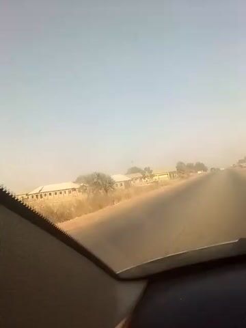 The video shows a view from inside a car driving on an asphalt road. The sky is light blue, indicating daytime. The air appears hazy, possibly due to dust or pollution. The road stretches into the distance. On the left side of the road, there are low-lying buildings with light-colored roofs and several windows. Vegetation, including trees and dry shrubs, is visible alongside the buildings and the road. Other vehicles are visible on the road ahead. The dashboard of the car is visible in the foreground.