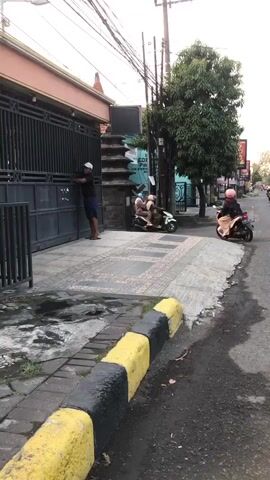 A person wearing a white cap and dark shirt is opening a black metal gate. A motorcycle is visible on the street to the right of the gate. The gate is located in front of a building with a tiled walkway leading up to it. The street is paved and has yellow and black curbs. Trees are present in the background.