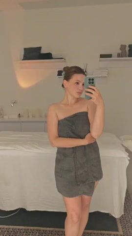 A young woman with blonde hair pulled up in a bun stands in a room, taking a mirror selfie with her phone. She is wrapped in a dark grey towel, with her left arm across her chest. The room appears to be a spa or treatment room. In the background, there is a white massage table covered with a sheet. White cabinets with various bottles and jars are visible behind the table, and shelves on the wall hold decorative items and what appear to be folded towels. The lighting is soft and warm, suggesting it might be evening. The woman has a neutral expression. The phone she is holding is a light blue color with a camera array on the back. The floor has a patterned rug.