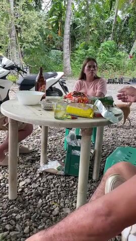 The video shows a group of people gathered around a white plastic table outdoors. A woman with long hair wearing a pink shirt is seated at the table and speaking. There is a bottle of beer, snacks, and glasses on the table. A small white and brown dog is visible in the background. The surroundings include lush greenery, trees, and a motorcycle parked nearby. The ground is covered with gravel. One man's legs are visible on the right side of the frame. The woman gestures with her hand as she speaks.