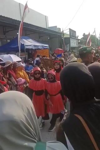 The video shows a street parade with many people walking in a line. In the foreground, several children are dressed in matching red outfits with feathered skirts and headbands. Behind them, other participants wear traditional conical hats and striped shirts. The parade is taking place outdoors on a street lined with buildings and tents. Several Indonesian flags are visible, suggesting a national celebration. A person in a military uniform is also present, observing the parade. The overall atmosphere appears festive.
