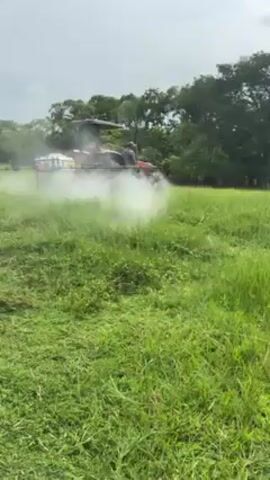 A man driving a red tractor with a spray rig attachment is seen moving through a field of green grass. The spray rig is emitting a white cloud of mist, presumably from a pesticide or fertilizer, as the tractor moves forward. The sky is overcast, and trees are visible in the background.