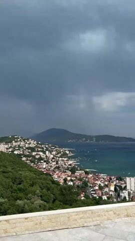 A high-angle, wide shot captures a sprawling coastal town nestled against a lush green hillside, with a dark, stormy sky looming overhead. The town, characterized by its dense cluster of white and red-roofed buildings, spills down towards a vibrant blue sea. Boats are dotted across the water, some anchored near the shore. In the foreground, a textured stone wall serves as a visual barrier, separating the viewer from the expansive landscape. The overall scene suggests a dramatic shift in weather, with the dark clouds and hint of rain creating a sense of foreboding or anticipation. The setting is identifiable as Sušćepan, Montenegro, adding a geographical context to the visual. The time of day is difficult to pinpoint precisely due to the overcast conditions, but it appears to be daytime. There is no visible text in the media.
