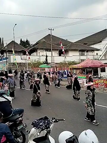 A group of people, mostly women, dressed in traditional clothing and hats are participating in a parade on a street. The individuals are walking in formation, some holding flags or banners. There are buildings and shops lining the street, and onlookers can be seen on the sides. The weather appears to be overcast. Motorcycles are parked in the foreground.