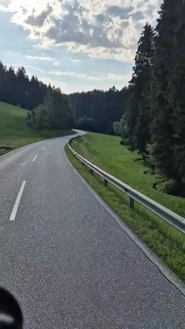 A first-person perspective video shows a paved road winding through a forested, hilly landscape. The road has a white dashed line marking the lane. On the right side of the road, there is a metal guardrail separating the road from a lush green grassy slope. Beyond the slope, a dense forest of dark green trees lines the horizon. The sky above is partly cloudy with a mix of white and gray clouds against a blue background. The overall scene depicts a scenic drive through a natural, wooded area during daylight hours.