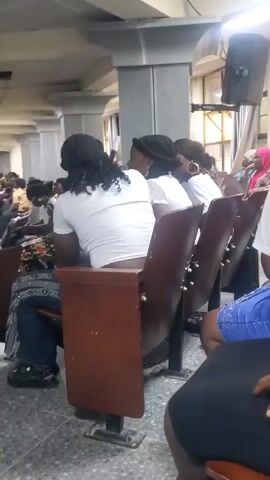The video shows a group of people sitting in chairs, possibly in a lecture hall or auditorium. The camera focuses on the back of a person wearing a white t-shirt and a black headscarf. To their right, several other people are visible, also seated and facing forward. Some are wearing white t-shirts and headscarves, while one person in the background is wearing a pink head covering. The chairs are made of wood and metal. The environment appears to be indoors, with gray tiled flooring and concrete pillars. A speaker is mounted on the wall in the upper right corner. The lighting is dim. The video is shaky, suggesting it was recorded handheld.