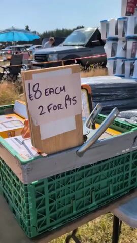 The video pans across an outdoor market stall on a sunny day. In the foreground, a green plastic crate holds several black, plastic-wrapped items. A sign on a wooden box reads "£8 EACH 2 FOR £15". To the right of the crate, a silver clamp is attached to a portable solar light. In the background, a dark SUV is parked, and people are milling around, with blue umbrellas and other market stalls visible. The overall impression is of a flea market or car boot sale.