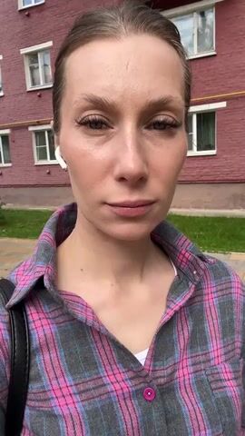 A medium shot of a woman with blonde hair pulled back, wearing white earbuds and a pink and grey plaid shirt over a white t-shirt. She is standing outside a brick building with windows. The woman is looking directly at the camera with a neutral expression. The background shows a sidewalk and green grass. The lighting is natural daylight.