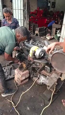 In what appears to be a cluttered workshop or garage, three men are gathered around a large diesel engine. One man, wearing a green shirt, is actively working on the engine, seemingly using tools to make adjustments or repairs. The engine rests on a wooden frame and is supported by a stack of bricks. Another man, wearing a striped shirt, stands nearby, observing the work in progress. A third man, partially visible, is holding a large, round metal component, possibly an air filter or muffler. The floor of the workspace is dirty and stained, with tools scattered around. In the background, there's more evidence of mechanical work, including a red machine and additional engine parts. Other individuals are faintly visible in the background as well, further implying this is a working garage.