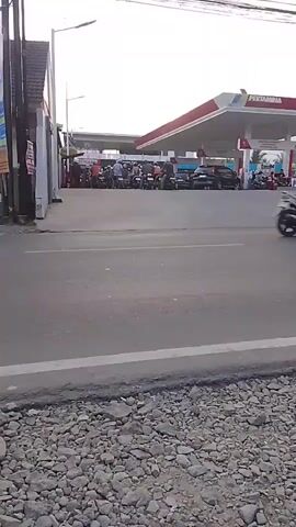 The video shows a street scene in front of a Pertamina gas station. A motorcycle with a rider passes by, blurred by motion. To the right, a white car drives past. In the background, there are other vehicles and people at the gas station. A sign for "Martabak Manis Mini D'Nikmat" with a phone number is visible on the right side of the frame. The road has a gravel shoulder.