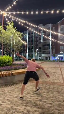 A young person with blond hair, wearing a pink t-shirt and black shorts, is captured mid-air performing a jump or flip. The background features a nighttime scene with string lights overhead and modern buildings with illuminated windows. In the foreground, a paved area with a textured surface is visible, along with a curved wooden bench and some purple flowers. The individual is positioned in the center of the frame, in motion.