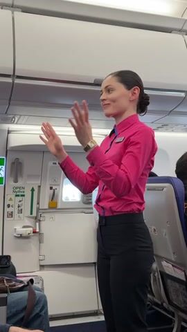 The video shows a young, light-skinned woman with her hair pulled back in a bun, wearing a pink button-down shirt with blue trim and dark pants, standing in the aisle of what appears to be an airplane. She is gesturing with her hands as if giving instructions or performing a demonstration. The emergency exit door is visible behind her, with a green sign indicating its operation. A passenger in a blue jacket can be seen seated in the background, and the leg of another passenger is visible in the foreground. The overall lighting inside the plane appears bright and even.