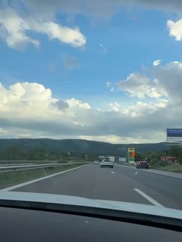 The video shows a view from the inside of a car driving on a highway. The sky is blue with scattered white clouds. In the distance, there are green hills. Several cars and a truck are visible on the highway ahead. There are also billboards on the right side of the road. The interior of the car, including the dashboard and part of the windshield, is visible in the foreground. The footage is shot from a stationary point of view within the moving vehicle.