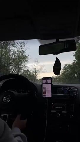 The camera is situated inside a dark car, positioned to capture the driver’s perspective. We see the steering wheel with the Volkswagen logo, the dashboard, and the rearview mirror reflecting the driver’s eyes. A smartphone is mounted in the center console, displaying what appears to be a music playlist. The car’s radio is tuned to Bluetooth audio.

Outside the windshield, rain is visible as the car moves along a road lined with trees. The overall tone is gloomy and overcast due to the rain and low lighting. The driver’s hands are lightly gripping the steering wheel.