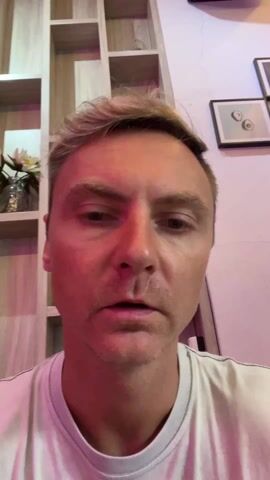 The video shows a close-up of a light-skinned man wearing a white t-shirt. He has short, light blonde hair and is looking directly at the camera, and points at his face. He looks to his right and then smiles at the camera. He then looks back up to his right, smiling wryly. Behind him is a shelving unit with a plant in a clear jar on one of the shelves. There is a framed picture on the wall behind him. The lighting has a pink hue.