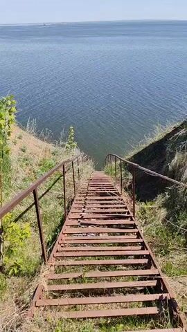 The video shows a view of a body of water from the top of a rusty, metal staircase. The staircase descends steeply towards the water, with a metal railing on either side. The surrounding terrain is grassy and slightly overgrown. The water appears calm, and the sky is blue with some wispy clouds. The overall impression is one of a somewhat dilapidated but scenic spot.