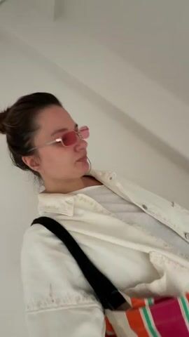The video shows a woman walking down a set of marble stairs. She is wearing rose-tinted sunglasses, a white T-shirt, a white jacket, and carrying a colorful striped tote bag with a black strap over her shoulder. The camera is positioned below her, angled upwards. Above the staircase is a window with small square panes.