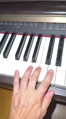 The video shows a close-up of a person's hand playing a digital piano. The hand is positioned over the keys, with fingers pressing down on several of them. The piano has black and white keys, and the person's hand is a light to medium skin tone. The top of the piano has buttons and labels such as "VOICE", "REVERB", "EFFECT", and "TOUCH". The background is blurred but appears to be a room with wooden furniture. The video is short, consisting of only a few seconds, and captures the motion of the hand as it plays the piano.