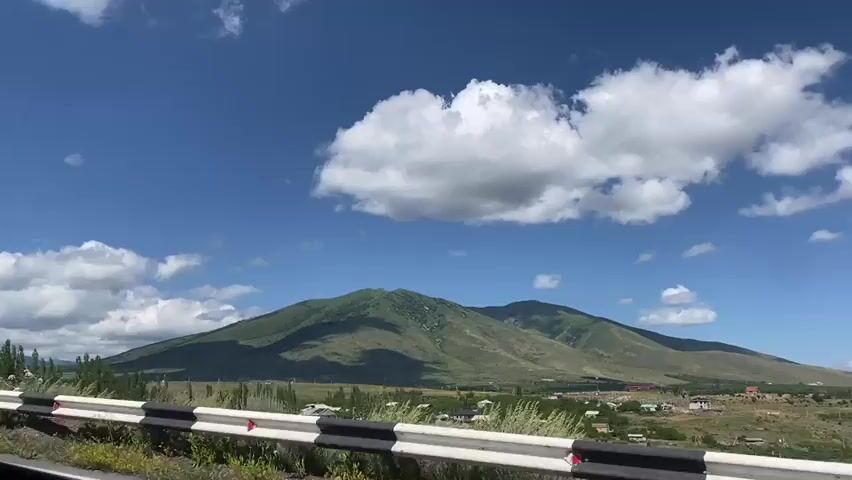 A stationary, wide shot captures a scenic landscape on a sunny day with fluffy white clouds against a bright blue sky. In the foreground, a reflective guardrail with alternating black and white stripes runs horizontally across the frame, marking the edge of a road. Beyond the guardrail, a grassy verge with wildflowers and scattered greenery leads to a small village nestled at the base of a majestic, green mountain range. The mountains, bathed in sunlight with areas of shadow, dominate the background. The village appears to consist of houses with red and brown roofs scattered amongst trees. A single road sign, a white triangle with a red border and a black arrow indicating a left curve, is visible on the right side of the frame, slightly behind the guardrail. The overall impression is one of a peaceful rural setting under a clear sky.