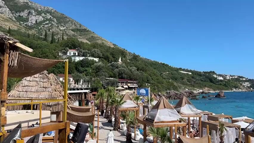 A wide shot captures a sun-drenched beach scene. In the foreground, a yellow railing runs horizontally across the bottom of the frame. Behind the railing, a collection of white and tan cabanas with draped white curtains and thatched roofs are set up on the pebbly beach. Palm trees are interspersed between the cabanas. Several people are seated or lounging in and around the cabanas. Beyond the beach, the turquoise water of the ocean stretches out, dotted with several boats and swimmers. In the background, a steep, tree-covered mountain slopes down to the sea, with white buildings visible on its side. The sky is a clear, vibrant blue.