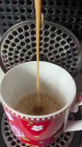 The video shows a close-up view of coffee being dispensed from a coffee maker into a red and white floral mug. The coffee is light brown and frothy as it fills the mug. The coffee maker has a black grid surface above the mug.
