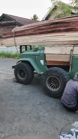 A heavily modified green utility vehicle, loaded with large wooden planks, is parked on a dirt or gravel surface. Three individuals are crouched down near the rear passenger-side wheel of the vehicle, appearing to be working on it. One person is wearing a gray t-shirt and a black baseball cap, another is wearing a brown t-shirt and light blue pants, and the third is wearing a black t-shirt. A black motorcycle with extensive decals is parked in the foreground, obscuring part of the scene. In the background, there are buildings, trees, and a large billboard advertising an event. The lighting suggests it is either early morning or late afternoon.