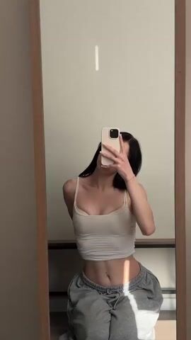 The video shows a young woman taking a selfie in a full-length mirror. She is wearing a white crop top and grey sweatpants. The setting appears to be a room with light-colored walls and a wooden floor. Sunlight is streaming in from the right, casting a strong shadow on her legs and the floor. The woman is kneeling and holding a smartphone in her right hand, which she uses to take the photos.

In the first frame, she is looking at the camera with a neutral expression. In the second frame, she pouts her lips. In the third frame, she closes her eyes and tilts her head back slightly. In the final frame, she opens her eyes and makes a peace sign with her left hand. The video appears to be taken during the daytime, given the sunlight. There is no visible text in the video. The woman's demeanor suggests a casual and playful mood. The overall impression is of a personal moment captured in a relaxed, indoor setting.