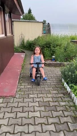A young girl with brown hair, wearing a blue and white star-patterned jumpsuit and blue slide sandals, sits on a black tricycle with a chrome fender. She is on a paved pathway made of interlocking grey pavers, lined with green plants and bushes. In the background, there is a brown-sided building with a white trim and a greenhouse. The scene appears to be outdoors during the daytime, possibly late afternoon or early evening given the soft lighting. The girl initially looks at the camera with a slight pout, then leans her head on the handlebars, smiling. She then looks back at the camera with a playful expression. The overall mood is casual and relaxed, suggesting a moment of playful interaction outdoors. There is no visible text in the video.