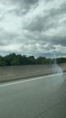 This is a video shot from a car, looking out the window at a cloudy sky and a landscape with wind turbines. The road is visible in the foreground, with a concrete barrier separating it from the rest of the scene. Green trees and bushes line the barrier, and in the background, several wind turbines are visible, their blades turning. The sky is filled with heavy, gray clouds, suggesting an overcast day, possibly with impending rain. A reflection of a person, likely the videographer, is visible in the car window. The video pans slightly as the car moves, showing a wider view of the wind farm and the sky.