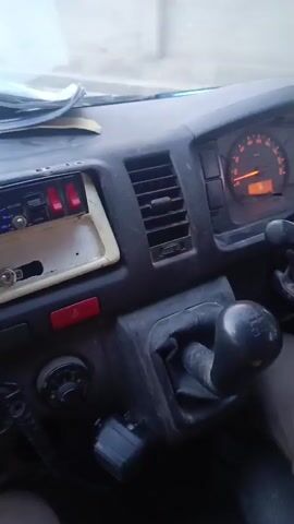 The video shows the interior of a vehicle, likely a van or truck, from the driver's perspective. The driver's hand is on the steering wheel. The dashboard is black with various controls and vents. A speedometer is visible, indicating a low speed or standstill. There is an aftermarket radio installed with blue lighting. A stack of papers rests on the dashboard. There is a glove compartment that is open with items inside. The driver appears to be wearing brown pants. The exterior is visible through the windshield, showing an indistinct urban or suburban environment.