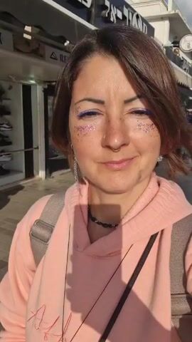 A woman is recording a selfie-style video on a sunny street. She has short, brown hair, fair skin with freckles, a septum piercing, and is wearing blue eyeliner and glitter makeup around her eyes. She is smiling slightly. She is wearing a light pink hooded sweatshirt and a black choker necklace, with a gray backpack over her shoulders.  Behind her is a tiled walkway with shops and buildings on either side. People walk in the background. Storefront signs visible include "SKECHERS" and "BIRKENSTOCK". The sky is partly cloudy. The woman does not speak or move much during the short video.
