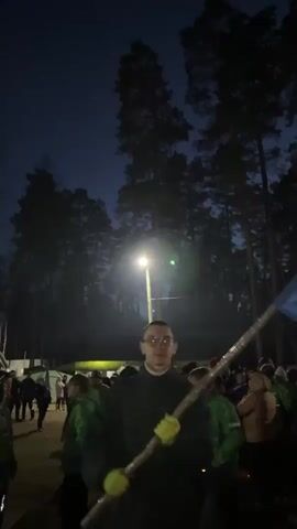 The video shows a man standing in front of a crowd of people, waving a flag. The flag is divided into three horizontal stripes of red, white, and blue, with a crest in the center. The man is wearing glasses, a dark green jacket, and yellow gloves. A light post illuminates the man and the crowd. The event is taking place at night, in what appears to be a park or forest. The background is dark and filled with tall trees. The camera angle is low, looking up at the man and the flag.