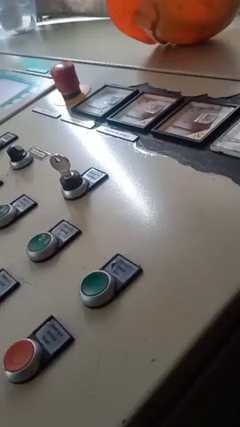 A close-up, slightly angled shot shows a control panel with multiple buttons, switches, and a keypad with a screen. The panel is light-colored and appears to be made of metal. There are rows of push buttons, some red and some green, each with a small label underneath. There are also several rotary switches with keys inserted into some of them. In the upper right corner of the frame, a portion of an orange object is visible, possibly a container or a piece of equipment. To the left of the control panel, a clear plastic container with liquid is visible, and behind it, a curtain or fabric is draped. The lighting suggests an indoor environment, possibly a workshop or industrial setting, with some glare reflecting off the smooth surface of the control panel. No people are visible in the video. The camera movement is minimal, providing a static view of the control panel.
