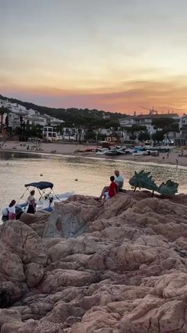 A vertical video clip opens on a rocky coastline with a bay and a town in the background. In the foreground, a group of people are gathered on the rocks. Two adults and a child sit on a large rock formation facing the water. To the left, several people stand near a small inflatable boat. In the middle ground, the bay is calm with several small boats anchored offshore. A sandy beach lines the bay, with more boats pulled up onto the shore. The town rises behind the beach, with white buildings and trees. The sky above is a soft gradient of orange, pink, and yellow, indicating sunset or sunrise. A green, stylized sculpture resembling a dragon's head is visible on the rocks in the foreground, near the seated individuals. The overall atmosphere is peaceful and scenic, suggesting a coastal vacation setting.