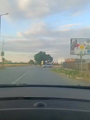 The video shows a view from inside a car, looking out through the windshield. The car appears to be in motion, driving on a paved road. In the distance, there is a silver minivan turning at an intersection. Beyond the intersection, there are trees and fields, with a cloudy sky overhead. There are also billboards visible on the side of the road. The time stamp indicates the video is 5 seconds long.