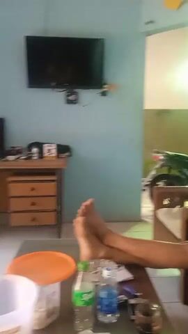 The video shows a man sitting on a wooden chair, looking at his phone. His legs are crossed, and he is wearing a black tank top and shorts. In the background, there is a TV mounted on the wall, a desk with a computer, and a doorway leading to an outdoor area where a motorcycle is visible. There are also bottles of drinks on a table in the foreground. The room has light blue walls.
