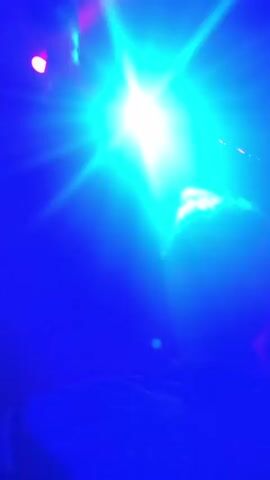 A video shows a group of friends in a crowded nightclub. The camera focuses on two friends, a woman with long dark hair and a man with curly hair, as they film themselves. The woman is smiling and shouting, and the man has a more neutral expression. The nightclub is lit with blue and purple lights, and there are many people dancing in the background. The camera moves around to show the crowd and the DJ booth. The music is loud and upbeat.