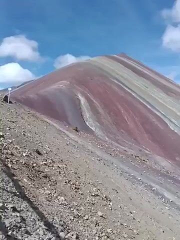 The video shows a colorful mountain range under a partly cloudy sky. The slopes of the mountains are striated with bands of red, gray, and other muted colors. The ground in the foreground is covered with rocks and gravel. In the distance, people can be seen at the foot of one of the mountains. There is also a dirt road winding through the landscape. The camera pans slowly across the scene.