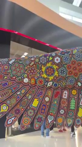 The video shows a large, ornate sculpture of an eagle, covered in colorful beadwork, inside what appears to be a shopping mall. The sculpture is very detailed, with intricate floral and geometric patterns covering the wings and body. In the background, there are people walking around and store signs visible, including a Puma store. The camera pans upwards, showcasing the full height of the eagle sculpture and the mall's ceiling. The overall setting is a well-lit, public space with the sculpture as the central point of interest.