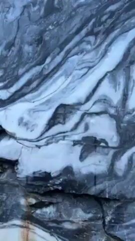 The video shows a close-up view of a large rock formation with swirling patterns of gray and white stone. The camera pans upward, revealing the height and extent of the rock face, with a few trees visible at the top. The texture of the rock is rough and uneven, with visible cracks and crevices. The overall environment appears natural, suggesting an outdoor location.