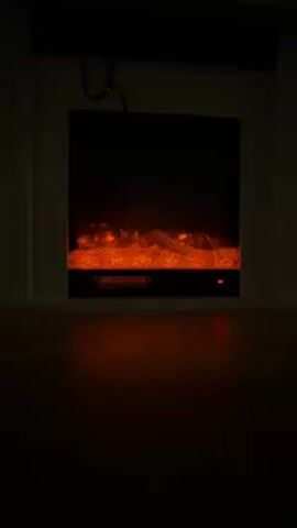 The video shows a stationary shot of an electric fireplace in a dark room. The fireplace is set within a white frame, and the faux flames and embers glow orange. A black cord hangs above the fireplace, partially obscured by the frame. The floor in front of the fireplace appears to be made of wood. The room is dimly lit, with the fireplace providing the primary source of illumination.
