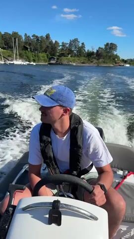 The video begins with a close-up of a young man wearing a blue baseball cap and a life vest, piloting a small inflatable boat. He is looking to his left, with a visible mustache. The boat is moving at speed, creating a white wake behind it. The background shows a body of water with trees and houses on the shore, and several sailboats docked in the distance. The sky is a clear blue with a few white clouds. The camera then pans out to show a wider view of the water, the shoreline, and the sky. The boat continues to move, and the wake is prominent. The overall scene depicts a leisurely boat ride on a sunny day.